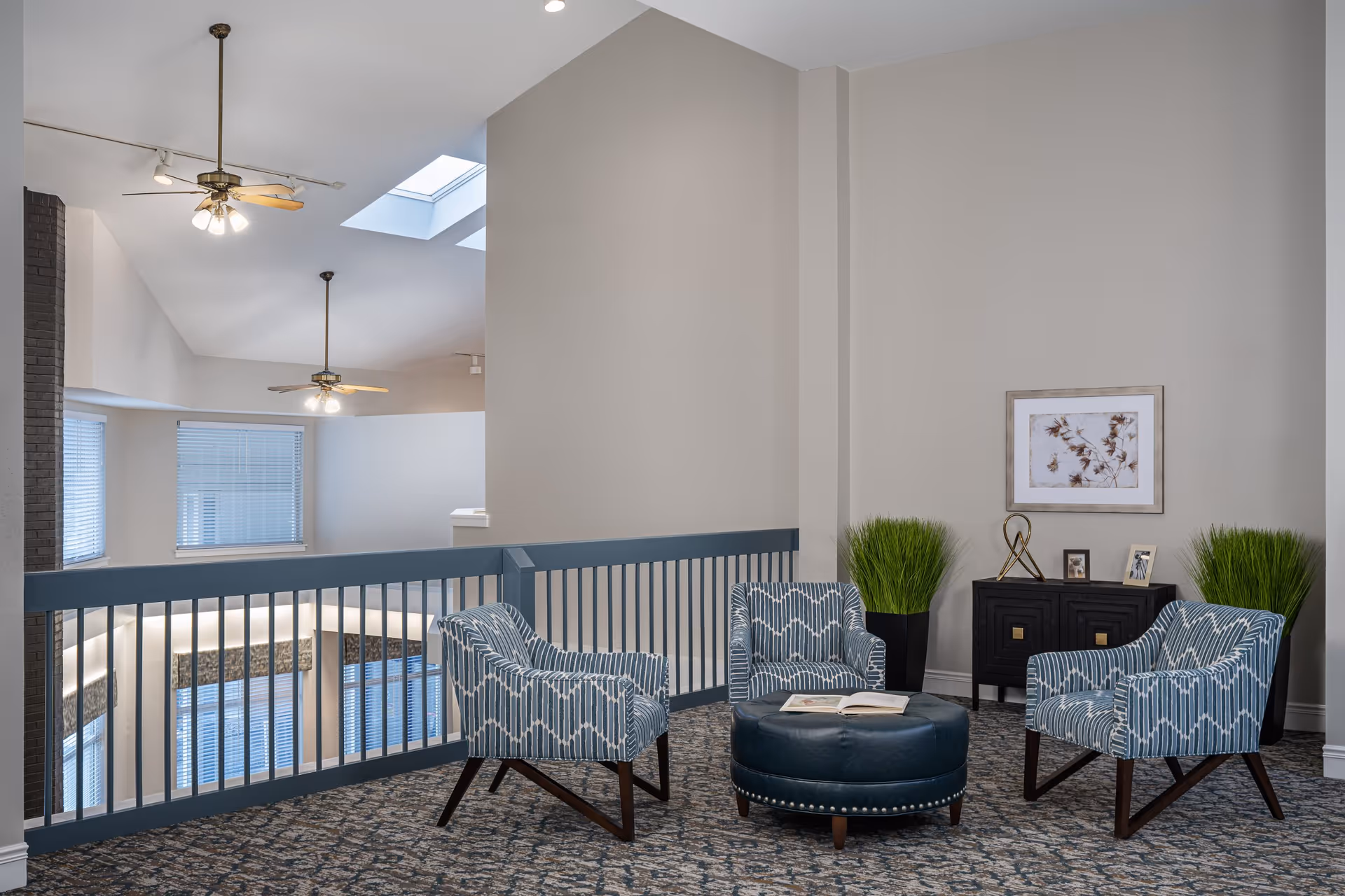 A cozy seating area in a senior living facility with three patterned blue armchairs arranged around a round blue ottoman with an open book on it. The space has a blue railing overlooking a lower level, two ceiling fans with lights, skylights, and a small black cabinet with decorative items and framed pictures. Two tall green plants flank the cabinet, and a framed artwork hangs on the wall above it.