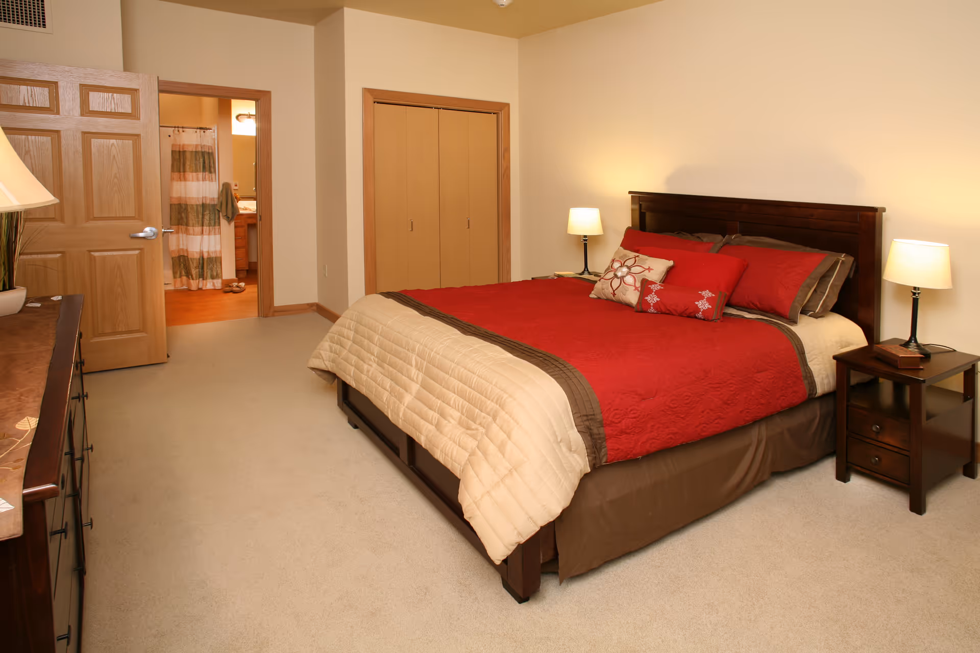 A bedroom with a large bed covered in a red and beige quilt, two bedside tables each with a lamp, a dresser with a lamp, a closet with double doors, and an open door leading to a bathroom with a shower curtain and towels.