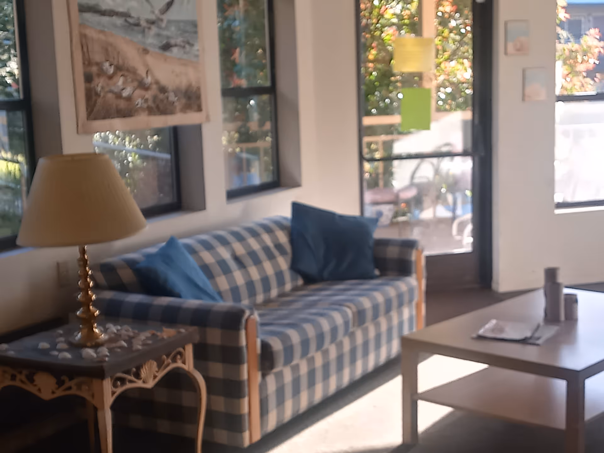 A cozy living room area with a blue and white checkered sofa adorned with two blue pillows. Next to the sofa is a decorative side table with a lamp and seashells on it. There are windows behind the sofa letting in natural light and a door leading to an outdoor patio with chairs and plants visible outside. A coffee table with some items on it is in front of the sofa.