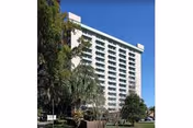 Tall white multi-story building with many windows, surrounded by trees and greenery under a clear blue sky.