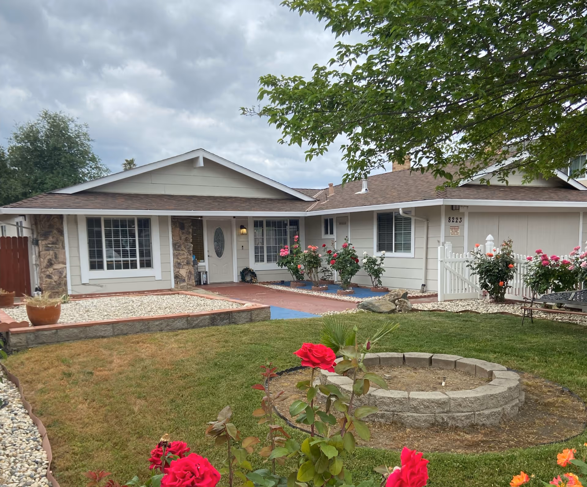 Front exterior view of a single-story residential building with a well-maintained garden featuring red and pink roses, a circular stone planter, and a white picket fence. The house has beige siding, stone accents around the windows, and a brown shingled roof under a cloudy sky.