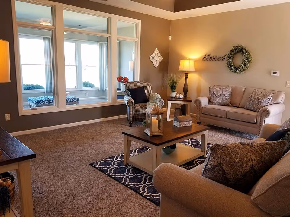 A cozy living room with beige walls and carpet, featuring a beige sofa with patterned pillows, a matching armchair, a checkered armchair with a dark pillow, a wooden coffee table with decorative items, a side table with a lamp and plants, and a large window looking into a sunroom with additional seating.