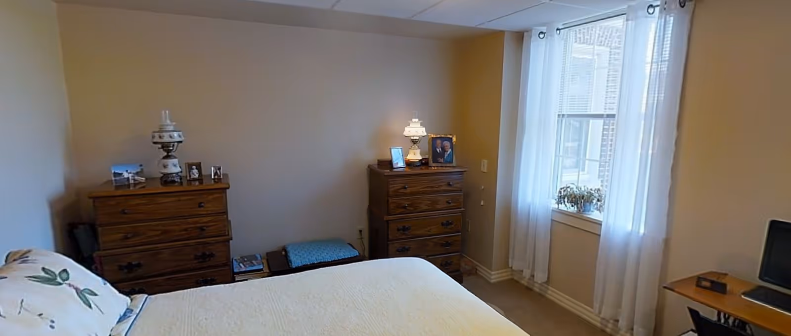 A cozy bedroom with a bed covered in a white quilt and a pillow with a floral design. There are two wooden dressers against the wall, each with decorative lamps and framed photos on top. A window with sheer white curtains lets in natural light, and a small desk with a computer monitor is visible on the right side.