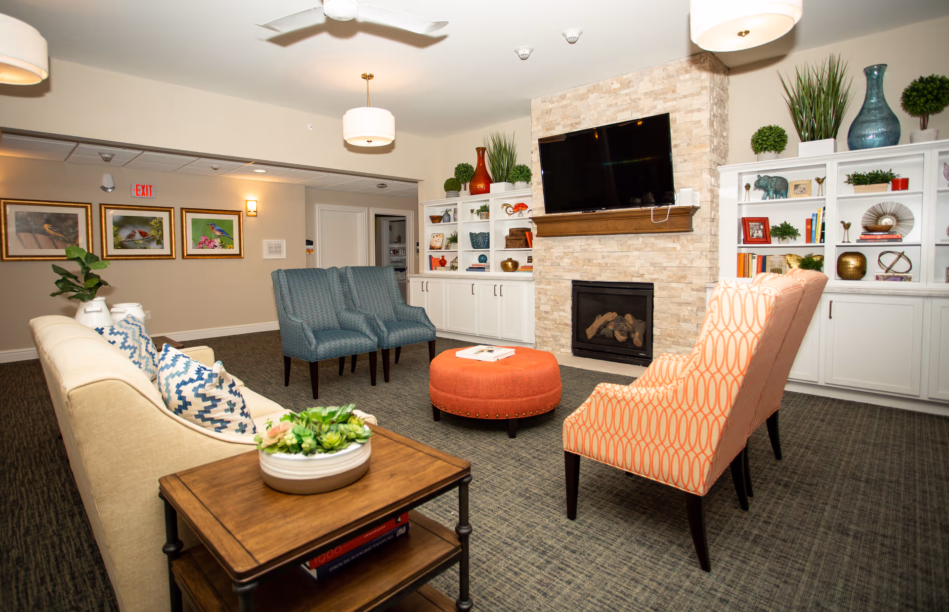 A cozy senior living facility common area with a beige sofa adorned with patterned pillows, two blue armchairs, and an orange patterned armchair arranged around a round orange ottoman. The room features a stone fireplace with a mounted flat-screen TV above it, white built-in shelves decorated with plants, vases, and books, and framed bird artwork on the wall near an exit sign.