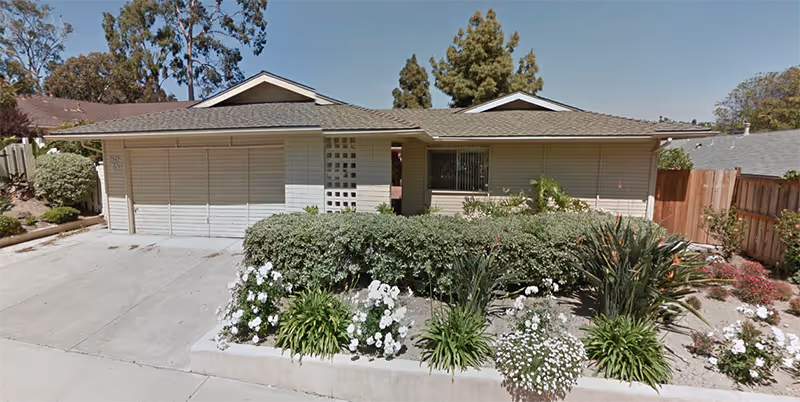 Front exterior view of a single-story residential building with a two-car garage, beige siding, and a small garden with green shrubs and white flowers in front. The sky is clear and blue.