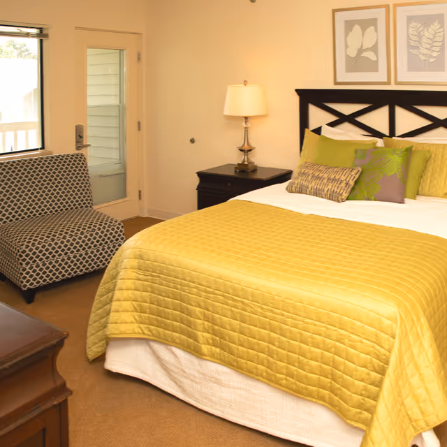 A cozy bedroom with a bed covered in a yellow quilt and multiple decorative pillows. There is a black nightstand with a lamp beside the bed, a patterned armchair near a window, and a door leading outside. Two framed botanical prints hang on the wall above the bed.