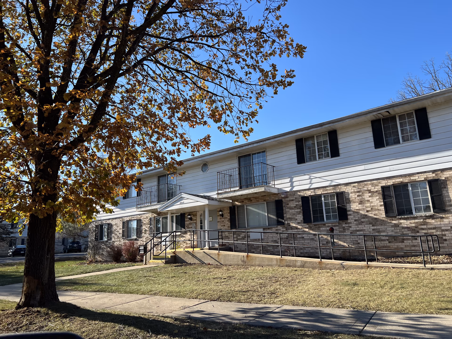 Exterior view of a two-story residential building with a brick and white siding facade, black window shutters, and a small balcony. There is a ramp leading to the entrance, a large tree with autumn leaves in the foreground, and a clear blue sky above.