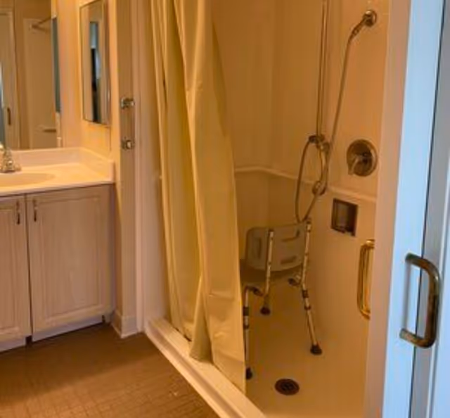 Bathroom with a shower area featuring a shower chair and a handheld showerhead, a yellow shower curtain, a sink with a cabinet below, and a mirror above the sink.