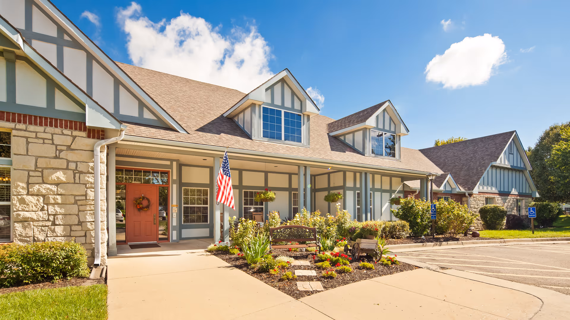 Exterior view of The Windsor of Lawrence senior living facility showing a large building with stone and wood paneling, a red front door with a wreath, an American flag, landscaped garden with flowers and bushes, and a parking area with handicap parking spaces under a blue sky with some clouds.