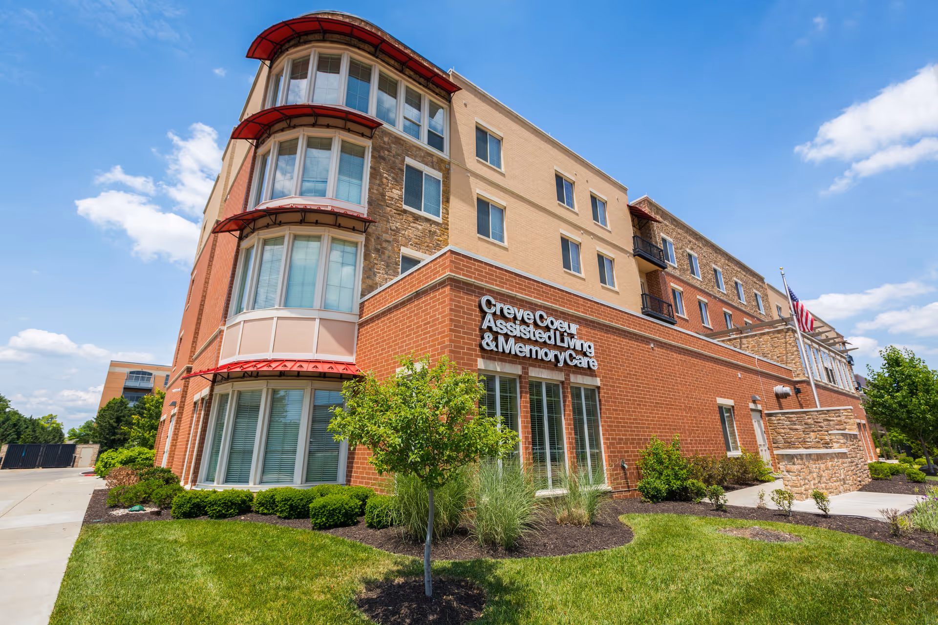 Exterior view of Creve Coeur Assisted Living and Memory Care building on a sunny day with blue sky and some clouds. The building features a combination of brick and stone facade with large windows and red awnings. There is a well-maintained lawn and landscaping with small trees and shrubs in front of the building.