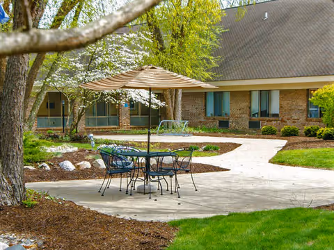 Outdoor patio area with a metal table and four chairs under a large striped umbrella, surrounded by trees, shrubs, and a building with multiple windows in the background.