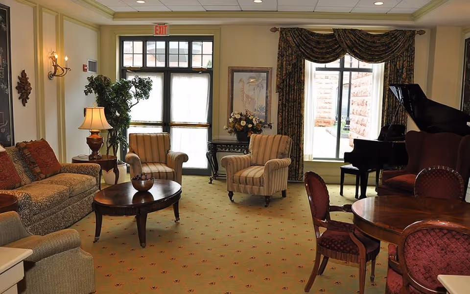 A cozy living room area with a patterned sofa, two striped armchairs, a round wooden coffee table, and a small round dining table with four red cushioned chairs. There is a grand piano near a large window with heavy drapes, a side table with a lamp, a potted plant, and a framed picture on the wall. The room has carpeted flooring and an exit door with glass panels.