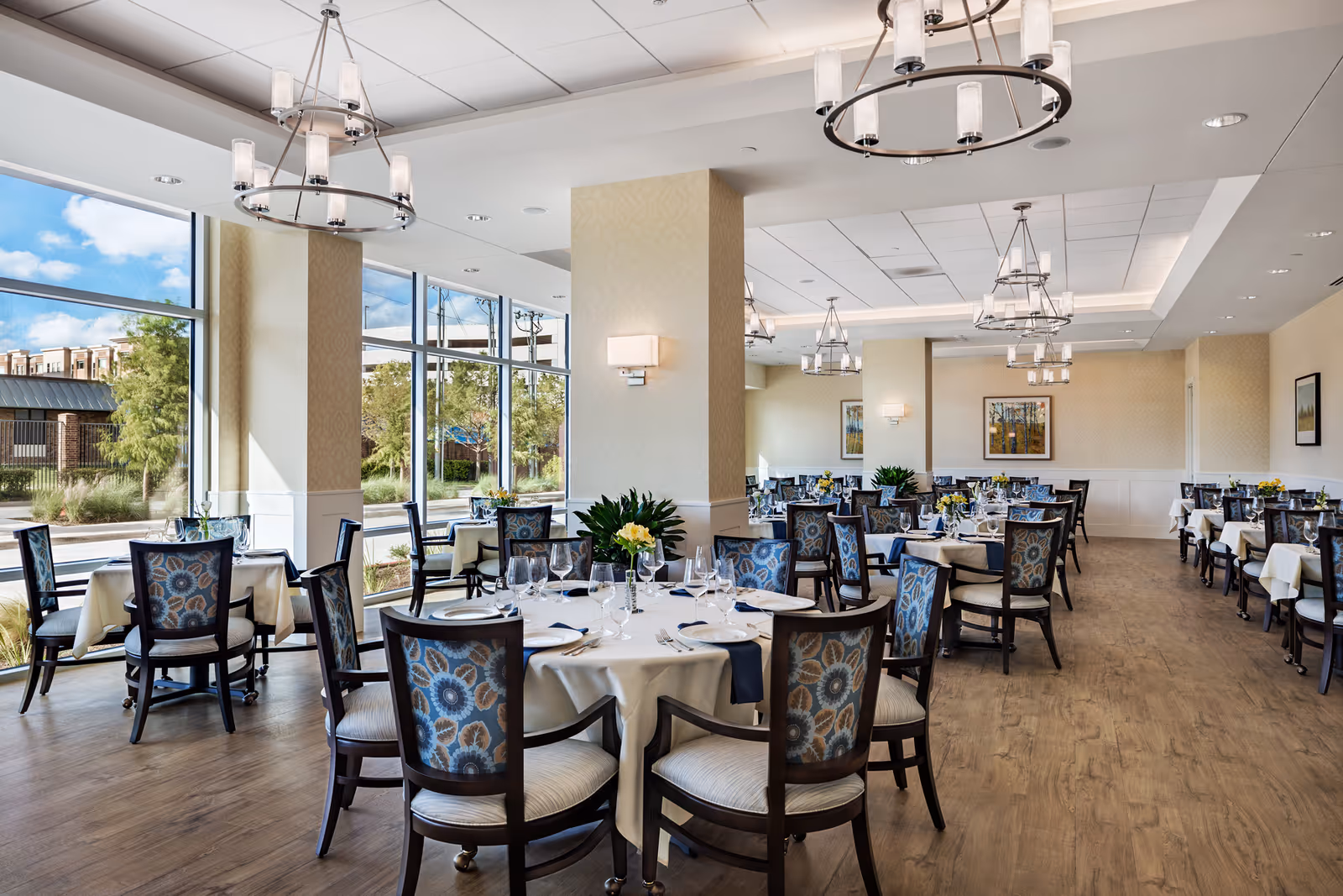 A spacious and elegant dining room with multiple round and rectangular tables covered with white tablecloths. Each table is set with plates, glasses, napkins, and floral centerpieces. The room features large floor-to-ceiling windows allowing natural light to fill the space, modern chandeliers hanging from the ceiling, and wooden flooring. The walls are light-colored with framed artwork and sconces.
