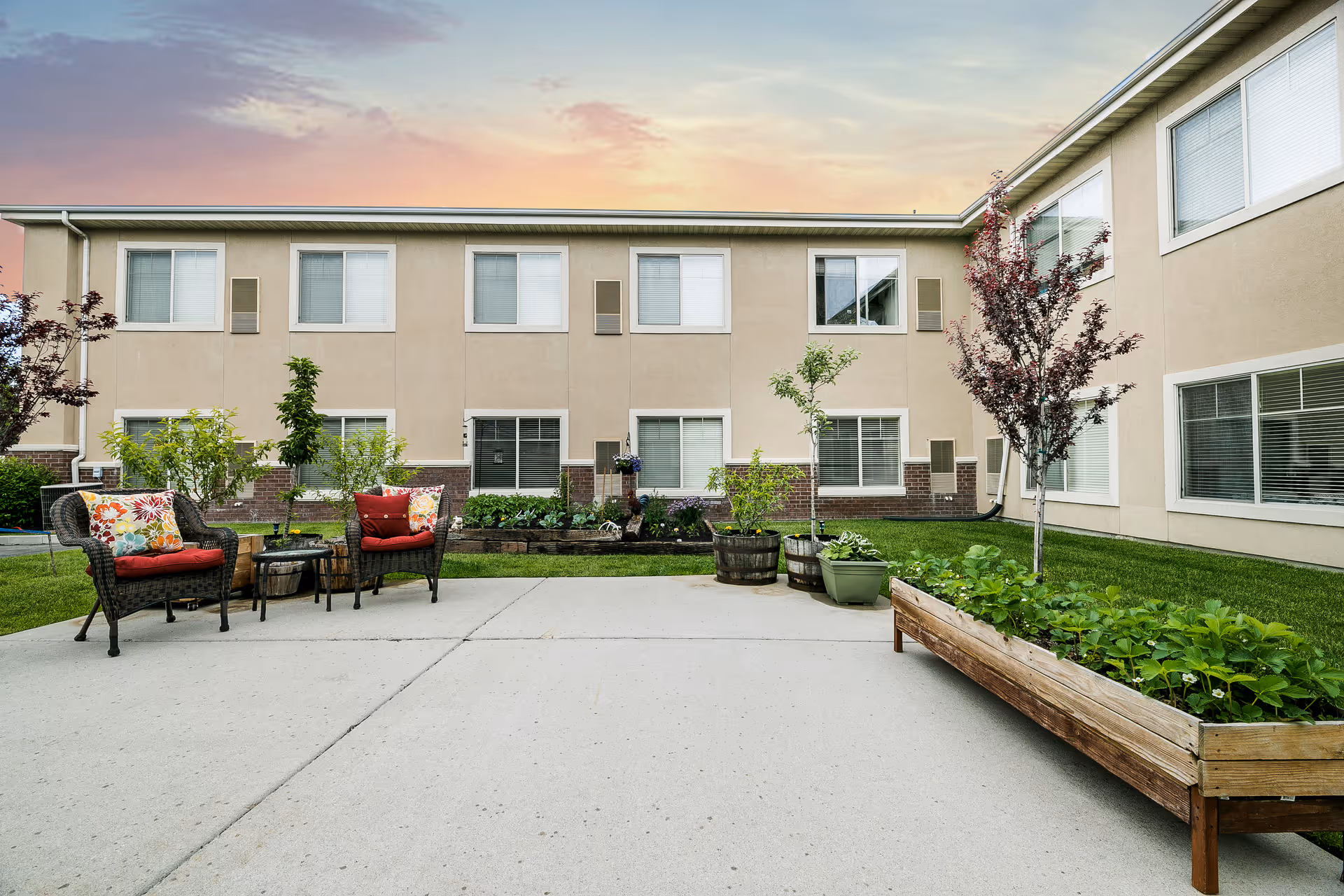 Outdoor courtyard area of a senior living facility with a concrete patio, two wicker chairs with colorful cushions, a small table, several potted plants, raised garden beds, and a two-story beige building with multiple windows in the background under a sunset sky.