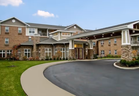 Exterior view of a senior living facility building with a covered entrance driveway, brick and stone facade, multiple windows, and a well-maintained lawn under a partly cloudy sky.