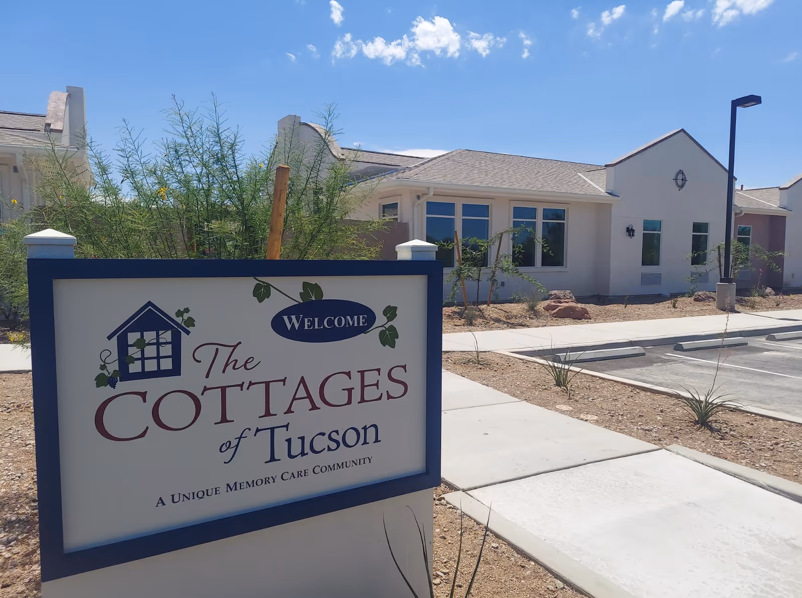 Outdoor view of The Cottages of Tucson facility sign with a single-story building and parking lot in the background under a clear blue sky.
