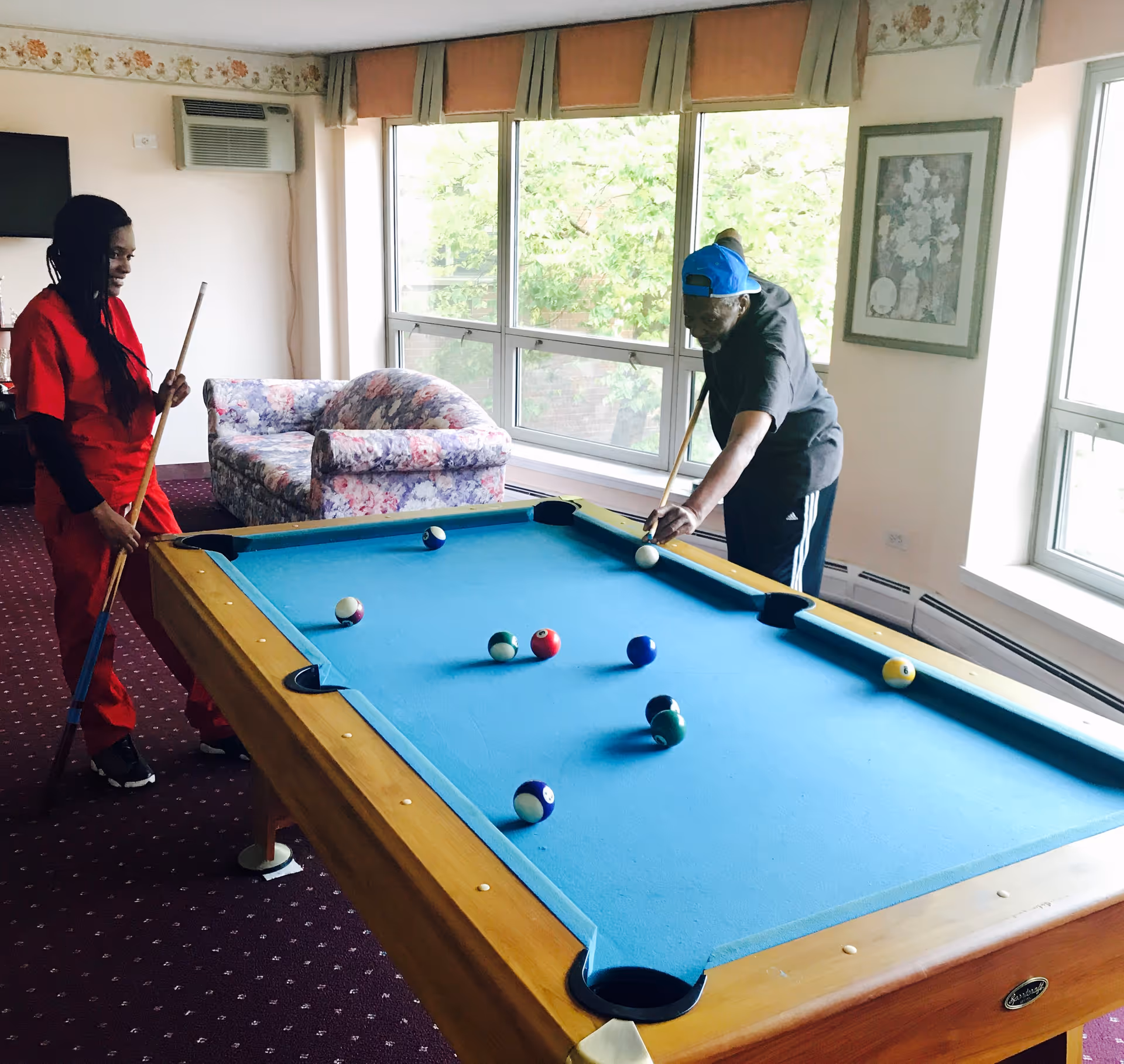 Two people playing pool in a room with large windows, a floral couch, and a framed picture on the wall. One person is preparing to take a shot while the other watches holding a pool cue.