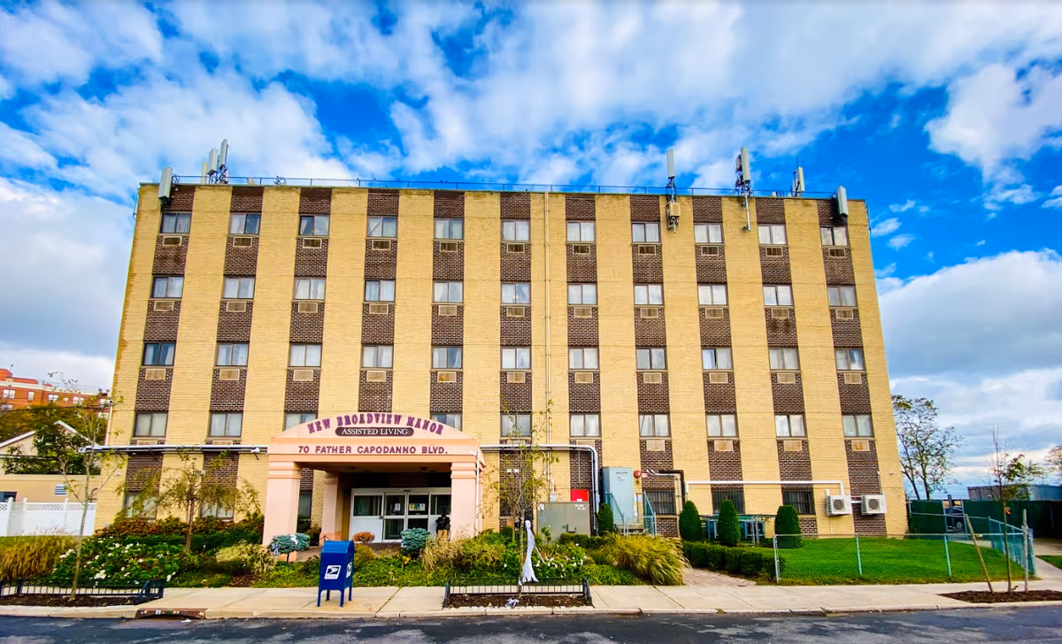 Front exterior view of New Broadview Manor, a multi-story assisted living building with a beige and brown brick facade, multiple windows, and a covered entrance with the address 70 Father Capodanno Blvd. The building is surrounded by a sidewalk, greenery, and a blue mailbox in front under a partly cloudy sky.