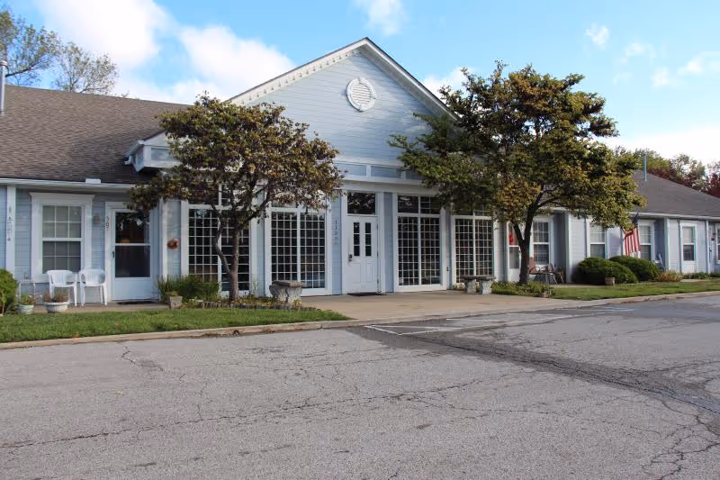 Exterior view of a single-story building with light blue siding and a dark roof, featuring large windows and a central entrance door. There are two trees and some shrubs in front of the building, along with a small paved area and a parking lot in the foreground. The sky is partly cloudy.