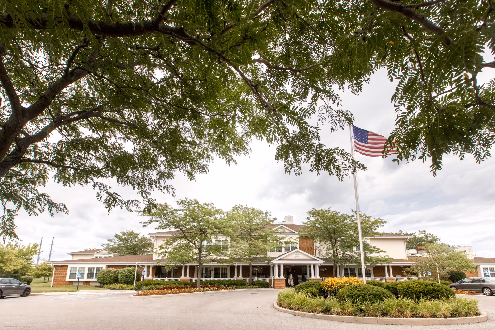 Front exterior view of Carriage Court Senior Living facility with a circular driveway, landscaped greenery, trees, and an American flag on a flagpole in front of the building under a cloudy sky.