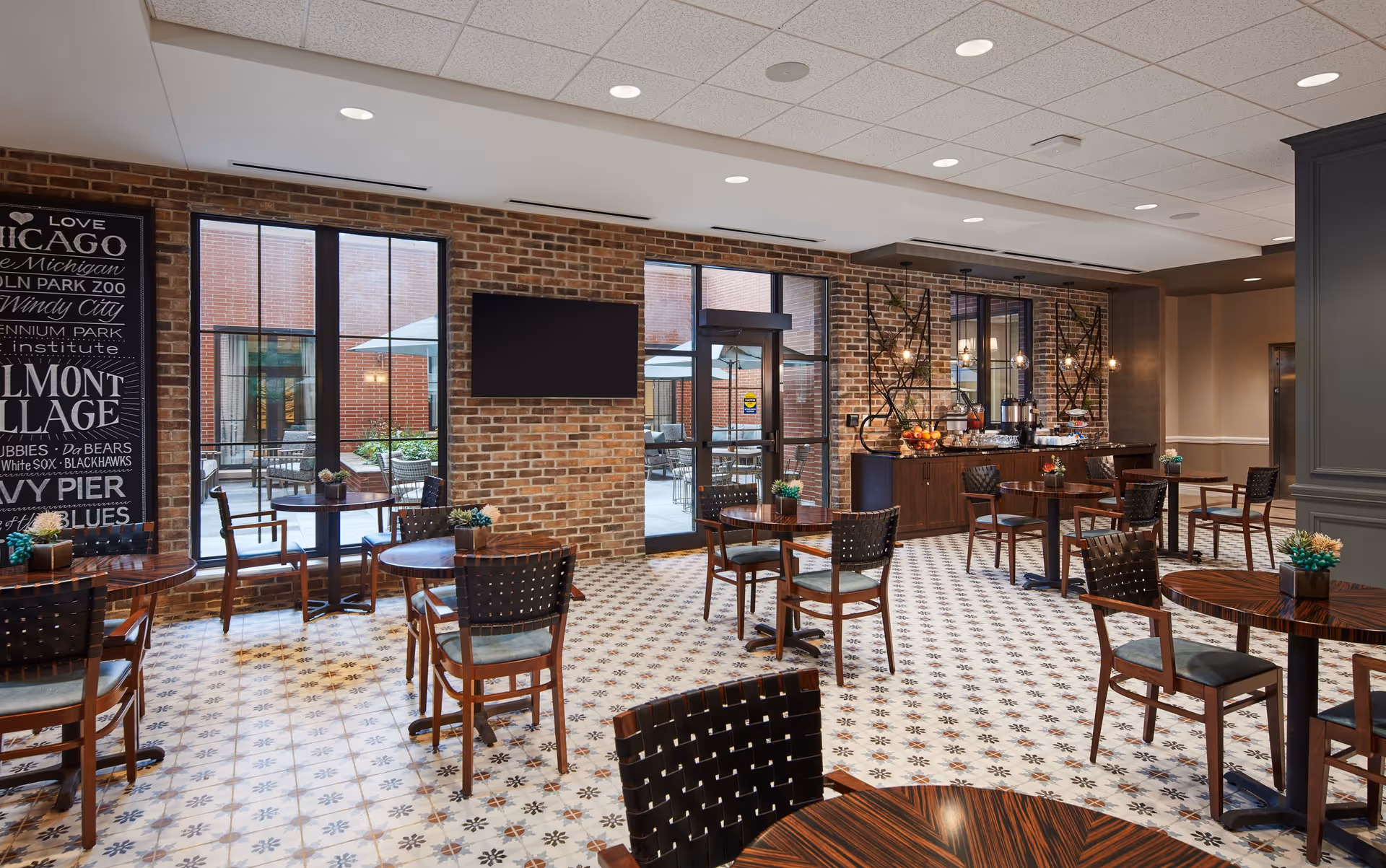 A bright dining area with patterned tile flooring, wooden tables and chairs with woven backs, and a brick wall featuring large windows and a door leading to an outdoor patio. There is a blackboard-style wall art on the left and a buffet counter with coffee and fruit on the right under hanging pendant lights.