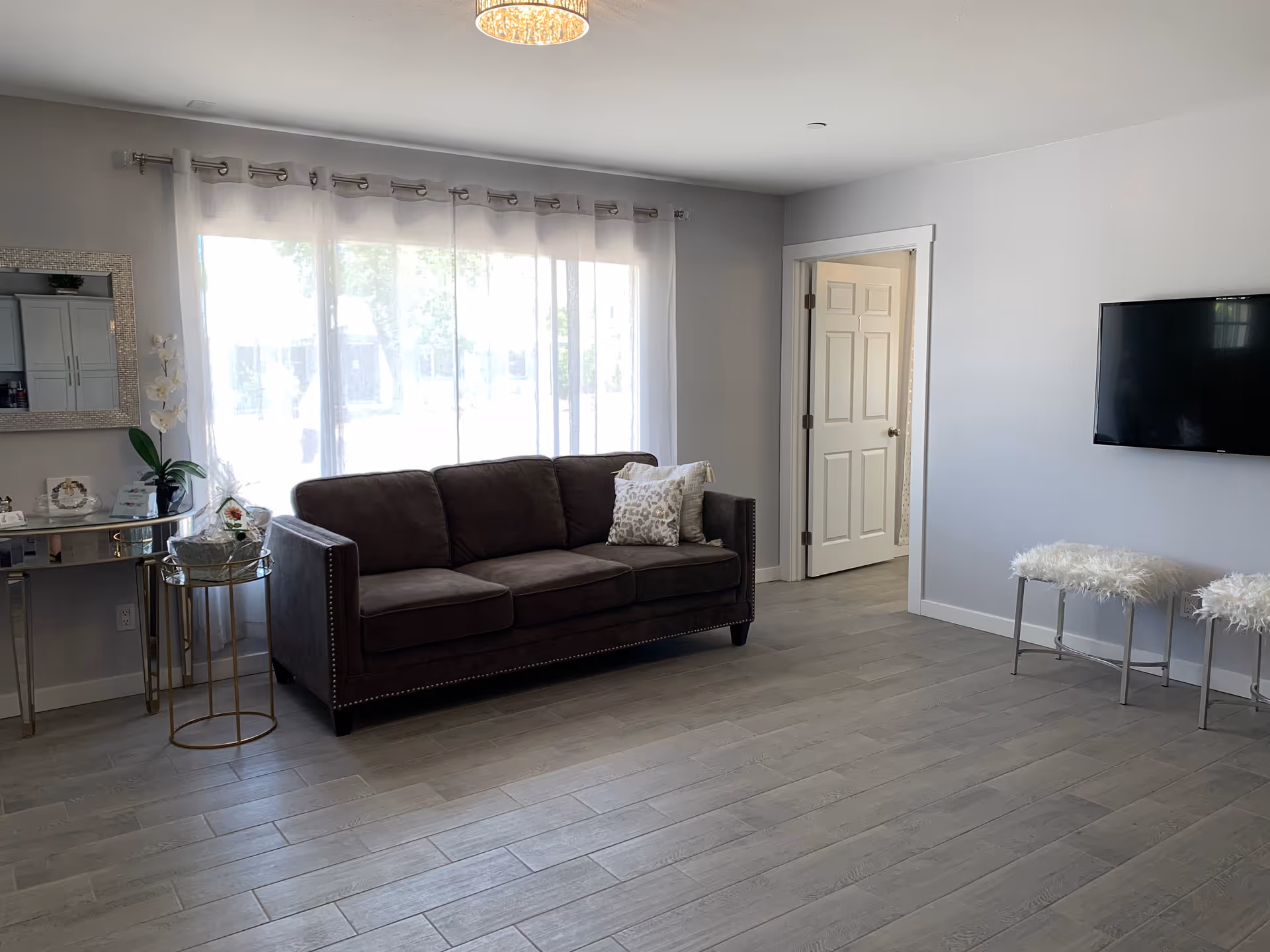 A bright living room with a large window covered by sheer white curtains. There is a dark brown sofa with a decorative pillow, a small round side table with a plant and decorative items, a wall-mounted flat screen TV, and two white furry stools. The room has light gray walls and wood-look tile flooring. A doorway leads to another room.