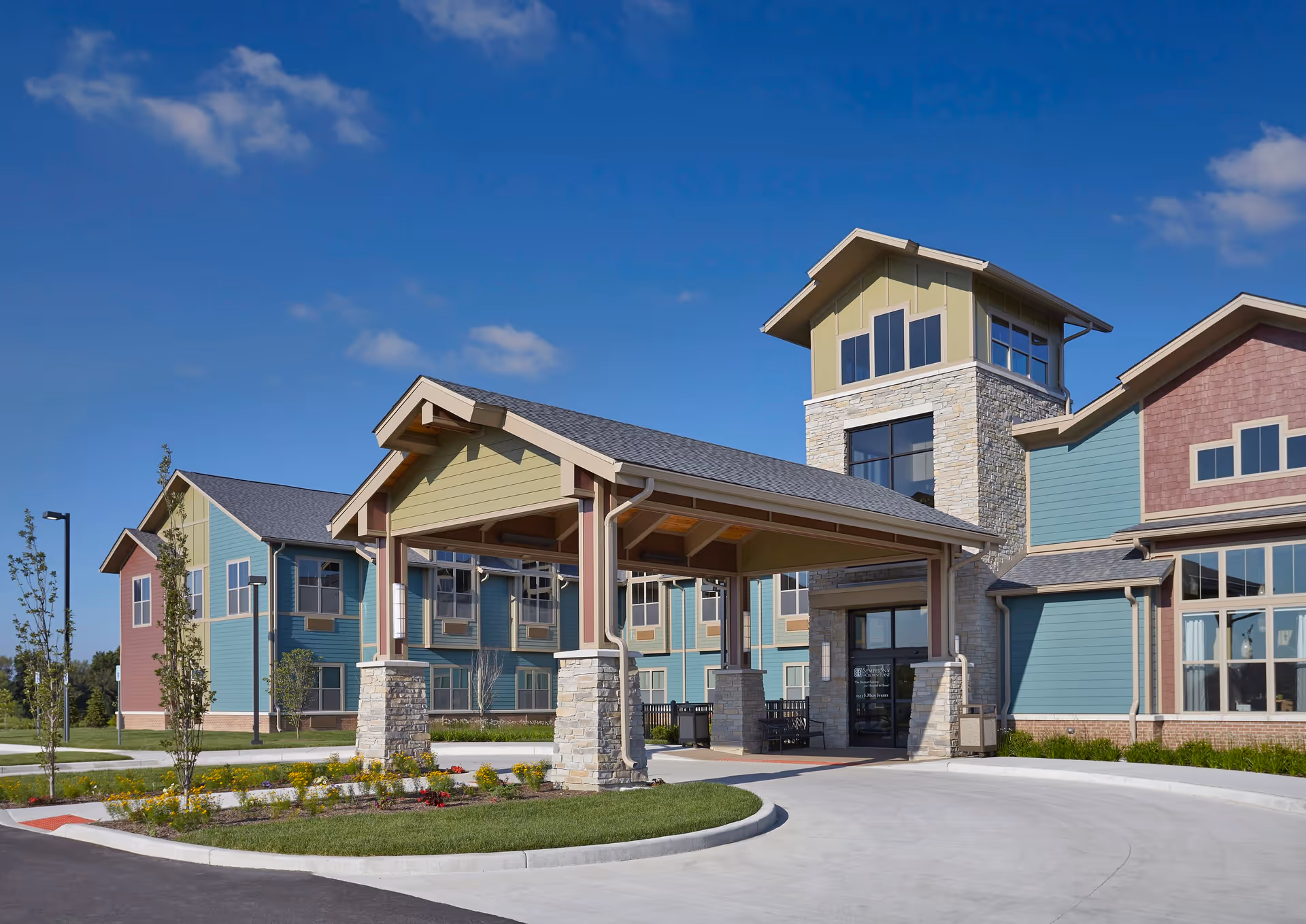 Exterior view of a senior living facility building with a covered entrance supported by stone pillars, colorful siding in shades of green, blue, and red, large windows, and a clear blue sky above.