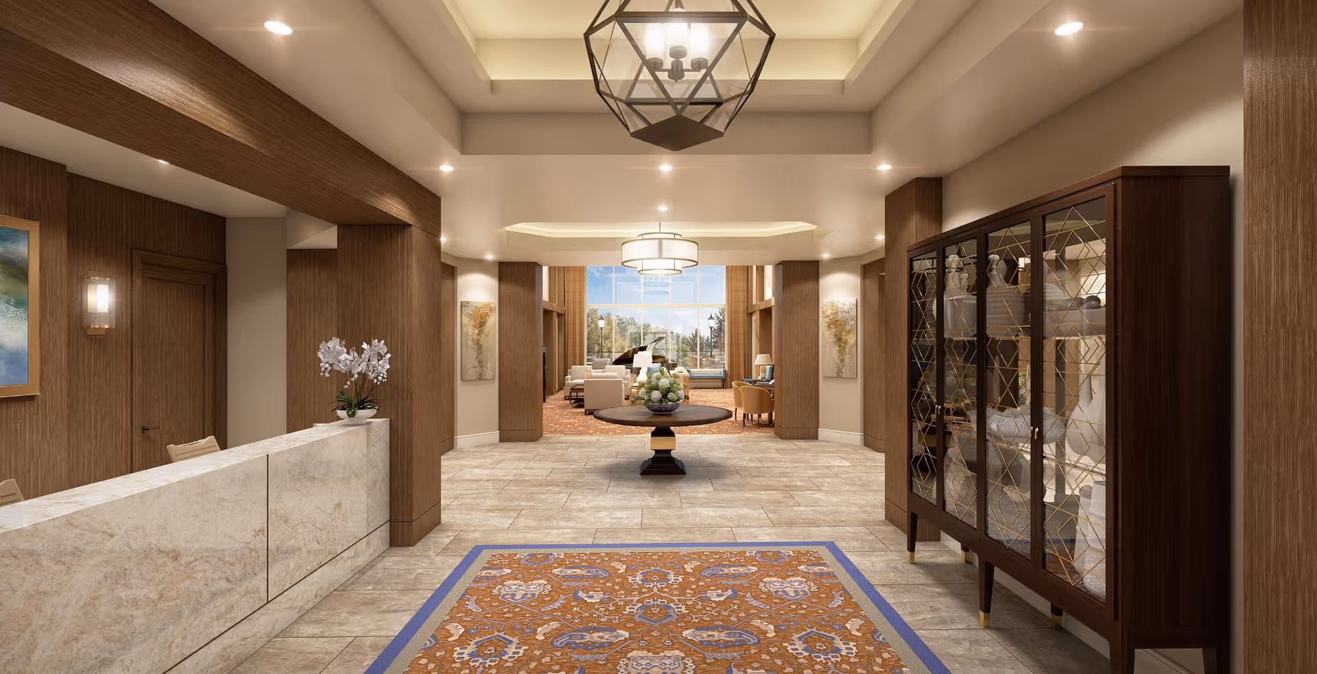A spacious and elegant interior lobby area with a marble reception desk on the left, a decorative wooden cabinet with glass doors on the right, and a round table with a floral centerpiece in the center. The room features warm lighting, wooden accents, a patterned rug on the tiled floor, and large windows at the far end letting in natural light.