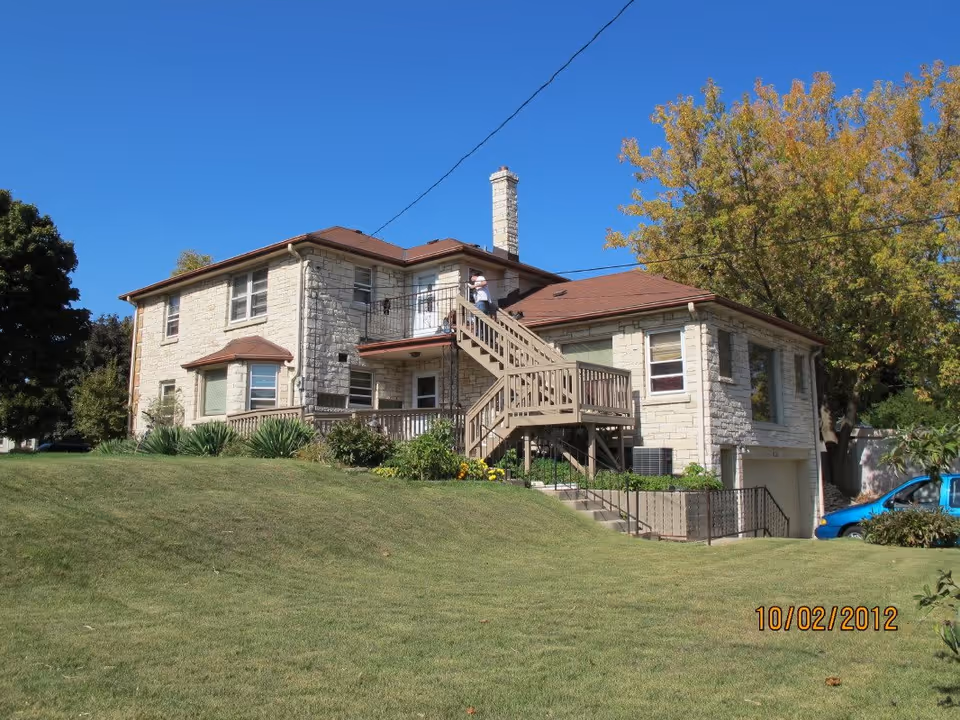 Exterior view of a two-story stone building with a brown roof, wooden staircase leading to the second floor, surrounded by green grass and trees under a clear blue sky.