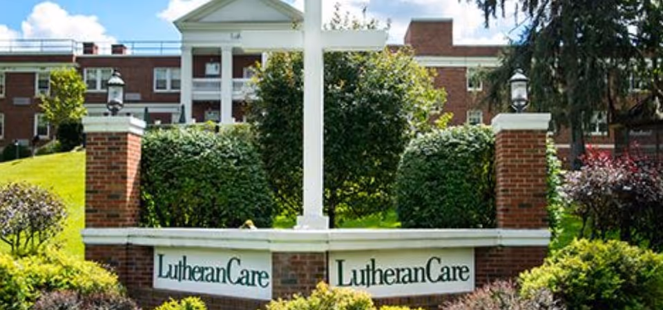 Outdoor view of the Lutheran Home of Central New York featuring a large white cross mounted on a brick structure with the words 'LutheranCare' displayed on two signs. The background shows a multi-story brick building with white columns and green bushes and trees surrounding the area under a partly cloudy sky.