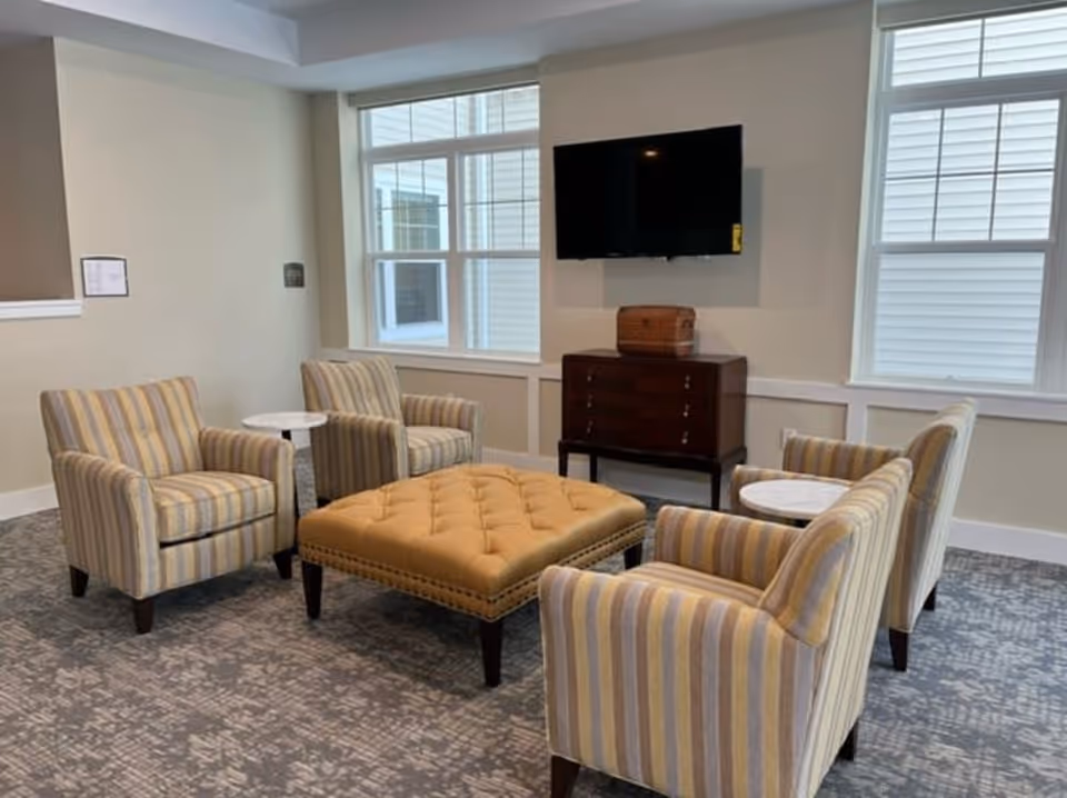 A cozy sitting area with four striped armchairs arranged around a large tufted ottoman. There are two small round side tables between the chairs. A flat-screen TV is mounted on the wall above a wooden cabinet. The room has two large windows letting in natural light and a patterned carpeted floor.