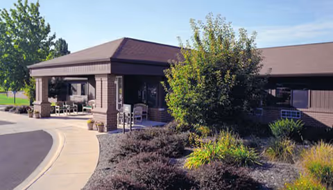 Exterior view of a single-story building with a covered entrance, surrounded by landscaped bushes and trees under a clear sky.