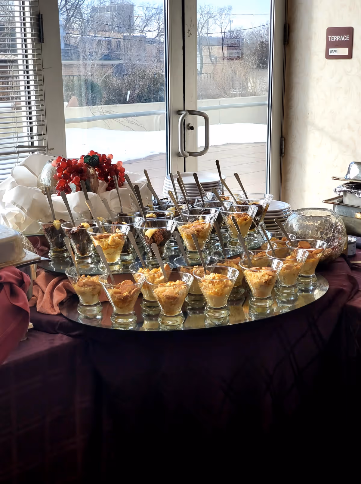 A table set with multiple glass dessert cups filled with various layered desserts, each with a spoon. The table is covered with a dark cloth and is positioned in front of glass doors leading to an outdoor terrace. There is a decorative arrangement of red berries and white cloth on the table, and a sign on the wall next to the door reads 'TERRACE OPEN'.