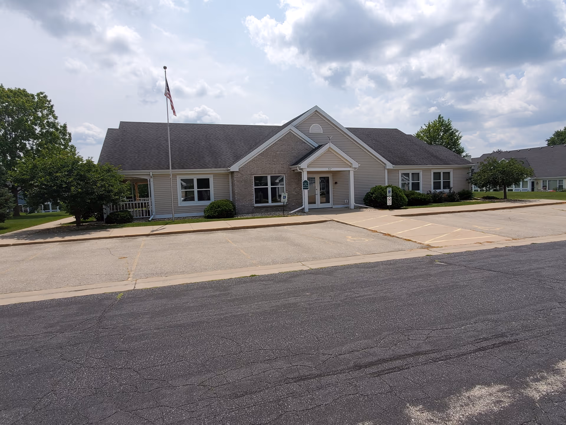 Single-story building with beige siding and a dark roof, featuring a small covered entrance with a white door. There is an American flag on a flagpole in front of the building, surrounded by green bushes and trees. The sky is partly cloudy and the building is situated next to a paved parking lot with marked spaces.