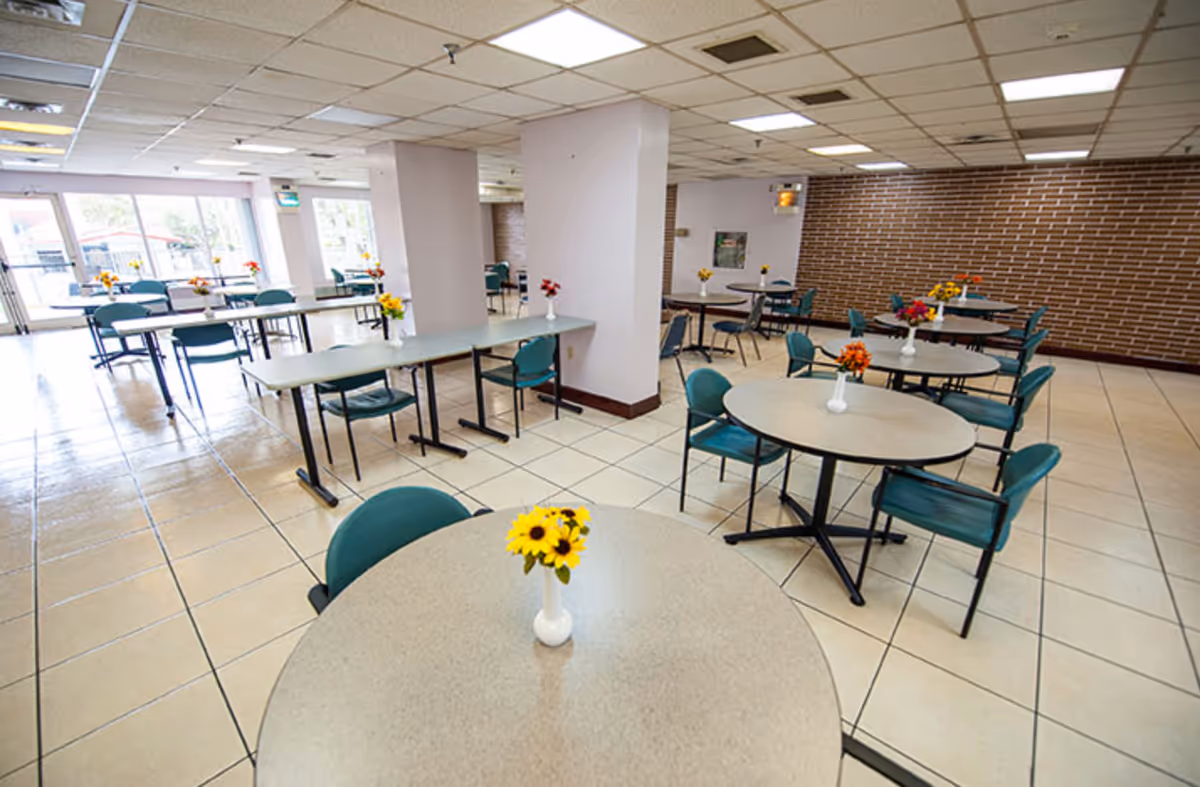 A spacious dining area with multiple round and rectangular tables, each surrounded by teal chairs. Small vases with colorful flowers are placed on each table. The room has tiled floors, a drop ceiling with fluorescent lighting, and large windows letting in natural light. A brick accent wall is visible on one side.