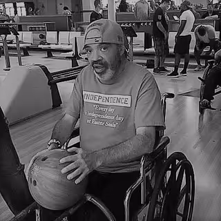 A man in a wheelchair holding a bowling ball at a bowling alley. He is wearing a backward cap and a t-shirt that reads 'INDEPENDENCE Every day Independence day is Easter Self'. In the background, other people are standing and preparing to bowl.