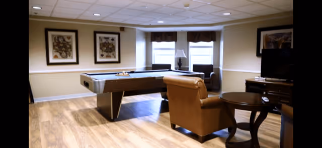 A spacious common area with a pool table in the center, a brown leather armchair, a round wooden side table, and a TV on a wooden stand. The room has light-colored walls, two framed artworks, two windows with valances, and a lamp between two armchairs near the windows.