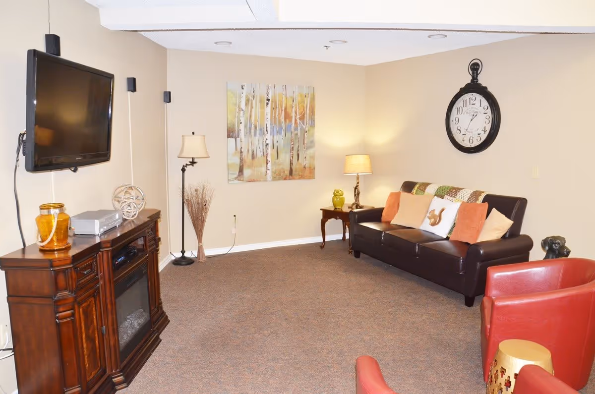 A cozy living room with beige walls and carpeted floor. The room features a dark brown leather sofa adorned with multiple cushions, a large wall clock above the sofa, a wooden side table with a lamp and a decorative owl, two red armchairs, and a small gold-colored side table. On the opposite wall, there is a mounted flat-screen TV above a wooden cabinet with decorative items and a floor lamp next to a wall painting of birch trees.
