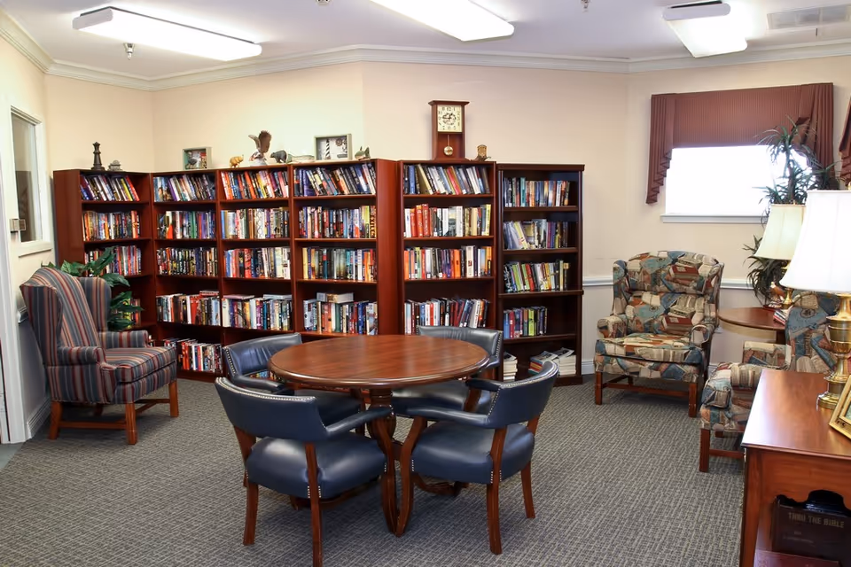 A cozy library room with several wooden bookshelves filled with books, a round wooden table surrounded by four blue cushioned chairs, and multiple upholstered armchairs with patterned fabric. The room has beige walls, a window with a valance, a clock on the wall, and a lamp on a wooden side table.