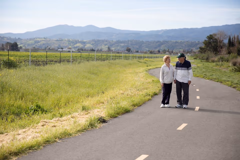 An elderly couple holding hands and walking on a paved path surrounded by green grass and fields with mountains in the background under a clear sky.