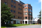 Exterior view of a multi-story brick building with several windows, a cross mounted on the wall, and a covered entrance area. There is a green lawn with trees in front and a clear blue sky above.