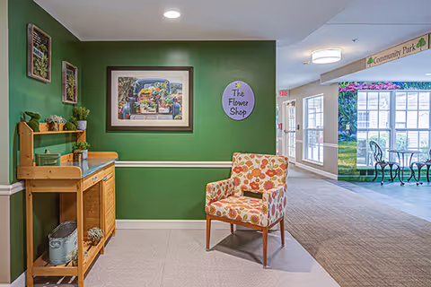 Interior view of a memory care community area with a green accent wall featuring framed artwork and a round sign that reads 'The Flower Shop'. A floral-patterned armchair is placed near a wooden shelf with decorative plants and items. In the background, there is a hallway leading to a bright area with large windows and a sign overhead that says 'Community Park'.