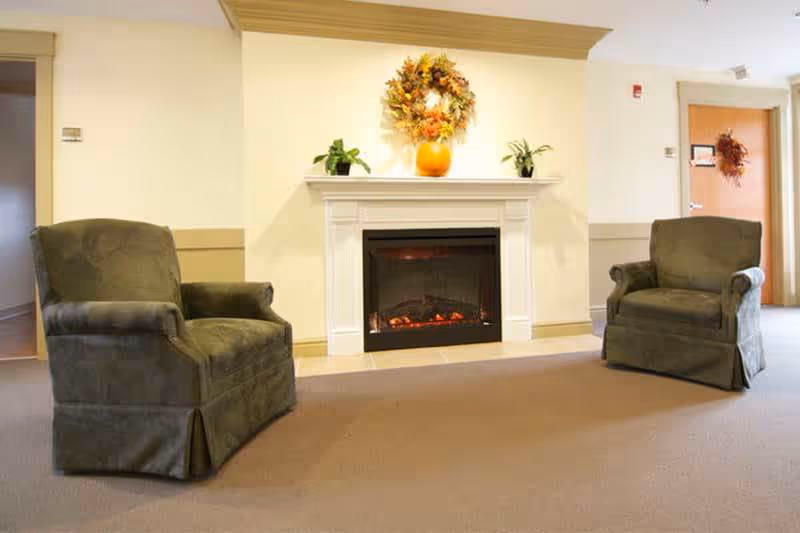 A cozy lobby area featuring two green armchairs facing a fireplace with a decorative mantle. A wreath and potted plants add a warm touch to the space.