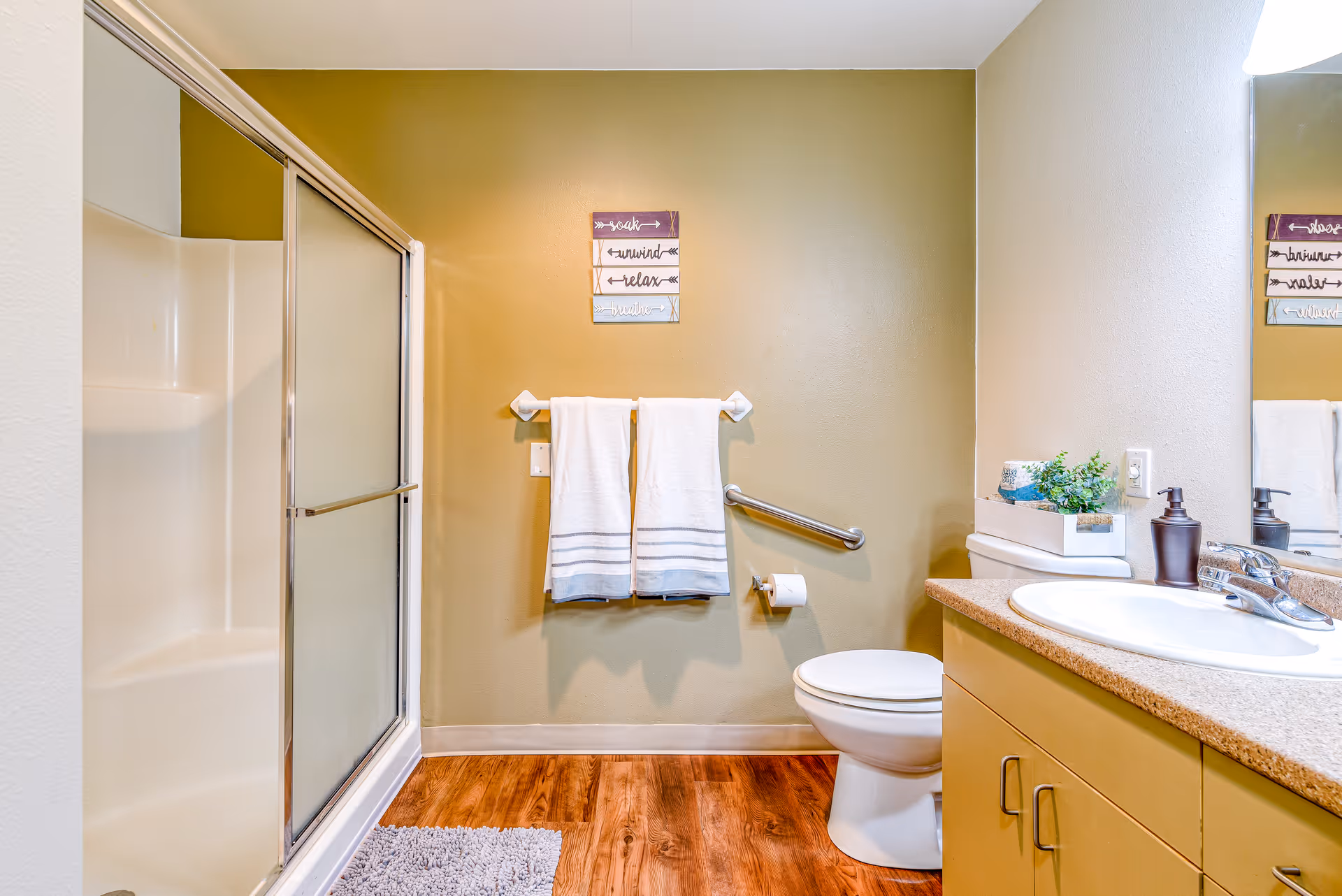 A bathroom with a walk-in shower with sliding glass doors on the left, two white towels hanging on a towel rack on a green wall, a toilet with a grab bar beside it, and a sink with a countertop and cabinet on the right. There is a small decorative sign above the towels and a small plant on the countertop next to a soap dispenser.