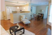 Open concept living space with a small seating area featuring a cushioned chair and ottoman, a coffee table with decorative items, a kitchen with white appliances and cabinetry, and a dining area with a wooden table and chairs. The room has hardwood floors and light-colored walls.