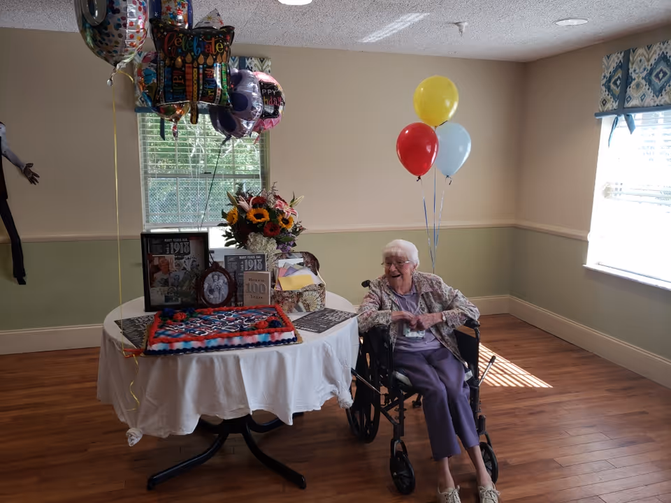 An elderly woman in a wheelchair smiling next to a round table covered with a white tablecloth. The table holds a decorated birthday cake, a bouquet of flowers, framed photos, and several colorful balloons. The room has wooden floors, light green and beige walls, and a window with patterned curtains letting in sunlight.