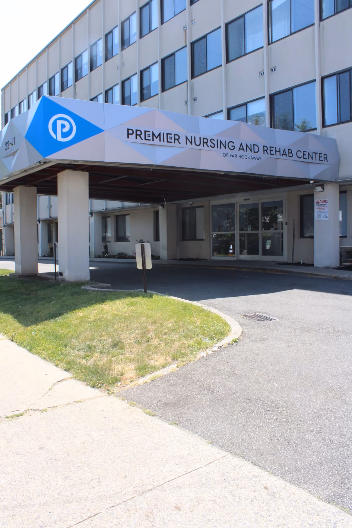 Entrance of Premier Nursing and Rehab Center of Far Rockaway showing a covered driveway with a sign above it displaying the facility's name. The building has multiple windows and a small grassy area beside the driveway.
