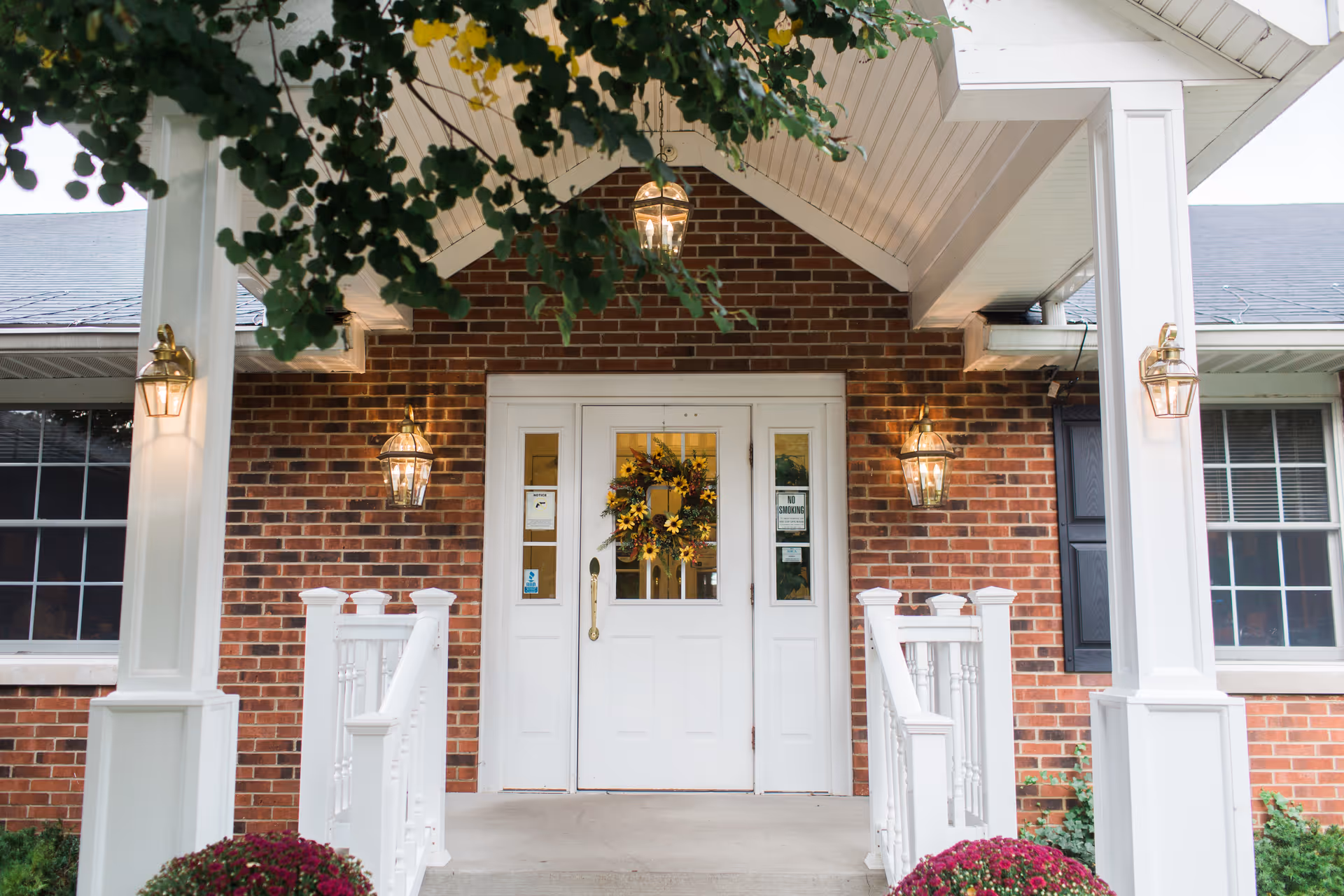 Front entrance of a brick building with white double doors decorated with a sunflower wreath, flanked by white railings and columns, with hanging lantern-style lights and a covered porch.