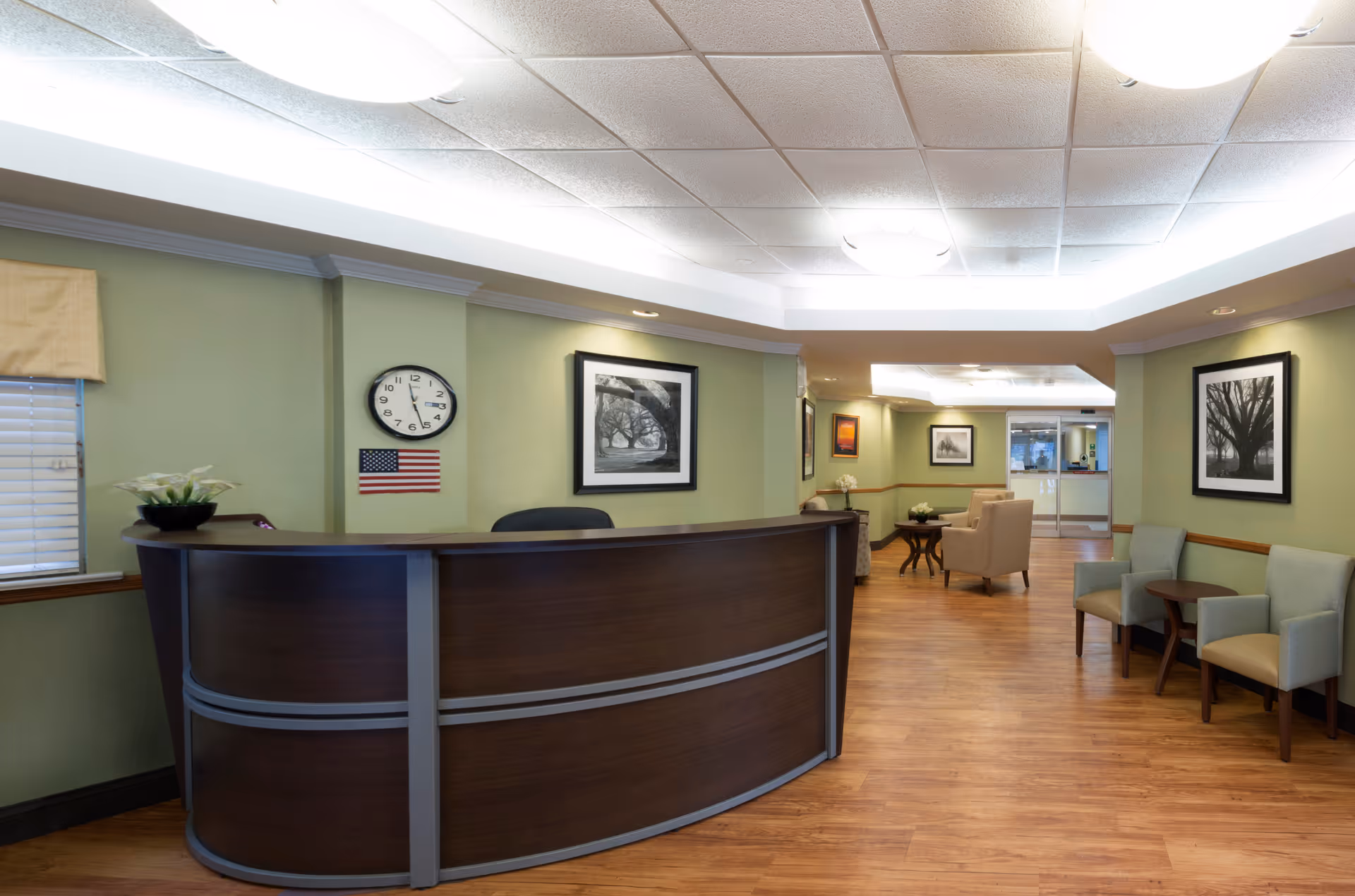 Reception area with a curved wooden desk, a clock showing 12:15, and an American flag on the wall. The room has light green walls, wooden flooring, several framed black and white photographs, and seating areas with chairs and small tables.