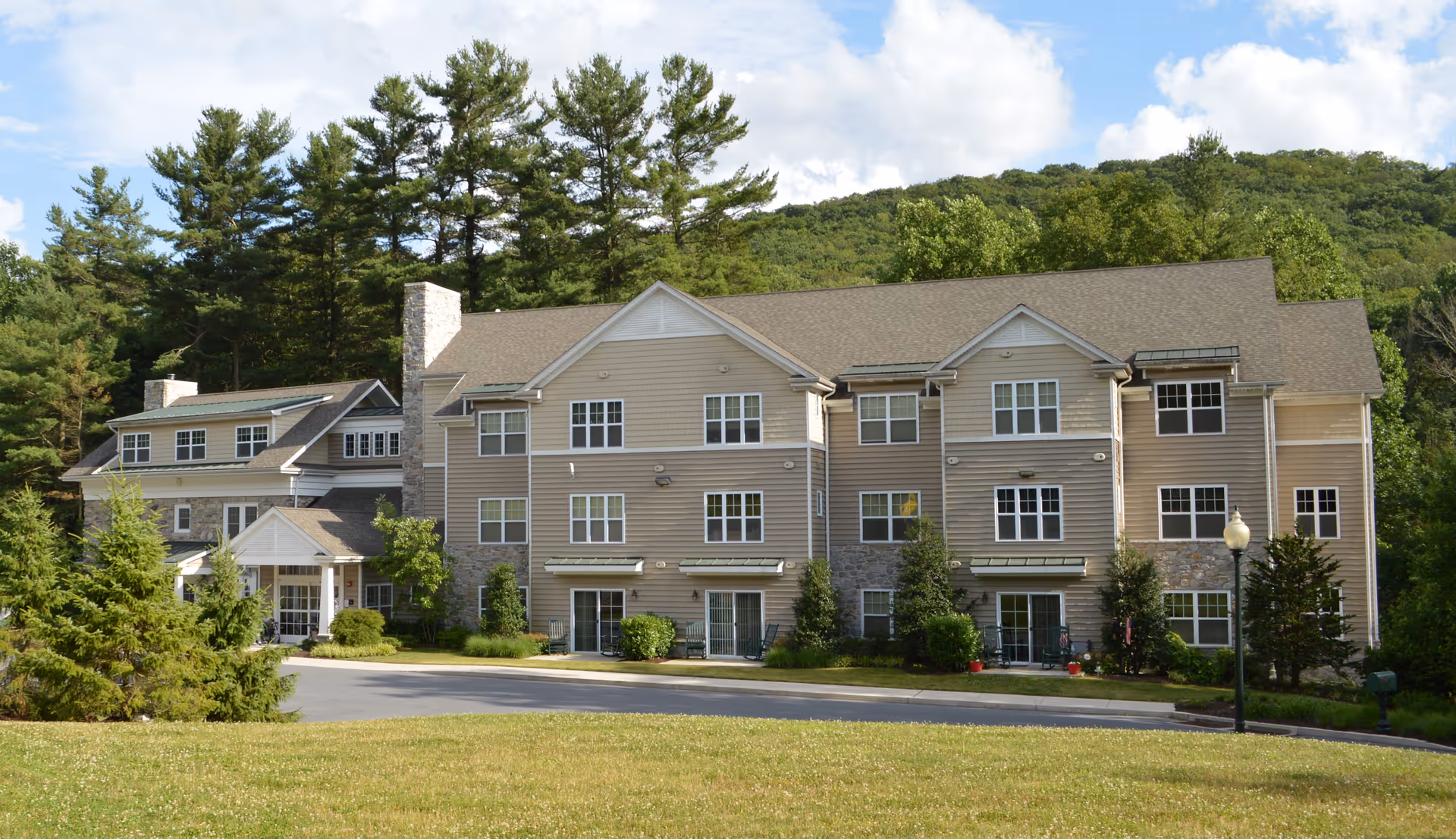 Exterior view of a multi-story senior living facility building surrounded by trees and greenery under a partly cloudy sky.